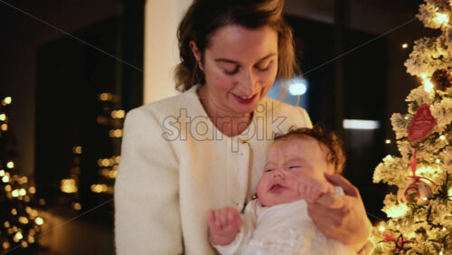 Video - Happy mother holding a baby indoors next to a decorated Christmas tree with warm lighting and soft background bokeh creating an intimate, festive atmosphere