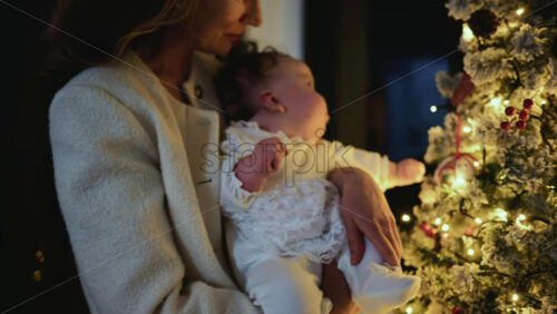 Video - A mother holding a baby, both dressed in white, while standing beside a decorated Christmas tree