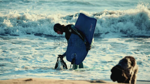 Video - Vallauris, France - December 27, 2025: Surfer in a wetsuit bending down on the shoreline to adjust fins while holding a surfboard