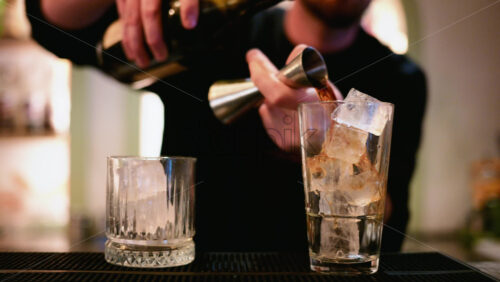 Video - A bartender pours a dark spirit from a metal jigger into a tall glass filled with clear ice cubes