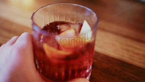 Video - Close up of a hand holding a glass filled with a red cocktail, ice cubes, and citrus peel