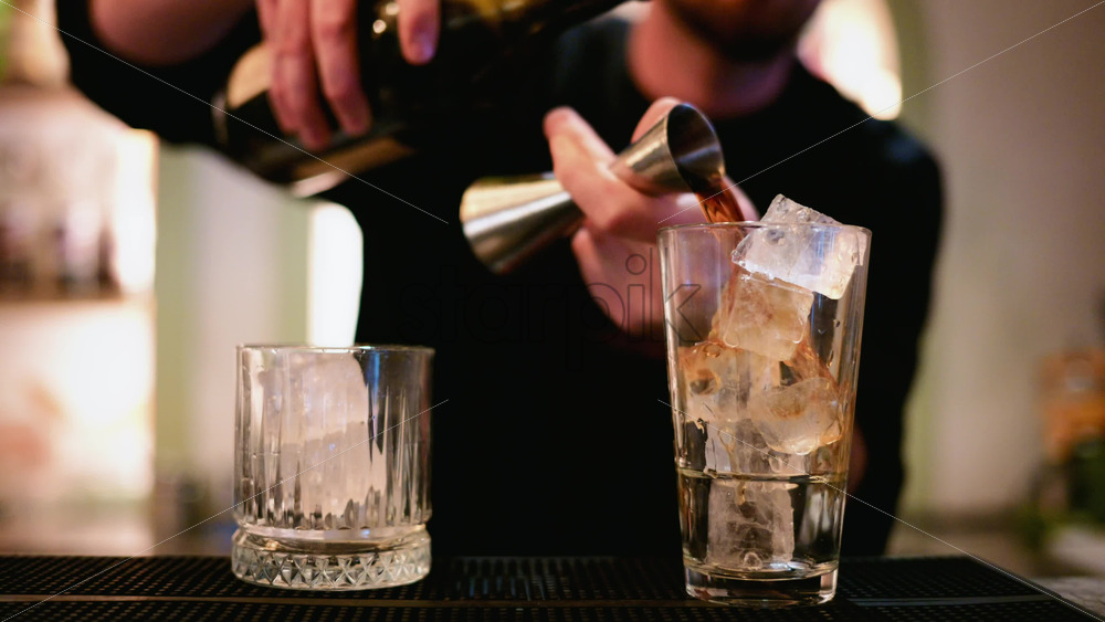 Video – A bartender pours a dark spirit from a metal jigger into a tall glass filled with clear ice cubes - Starpik Stock