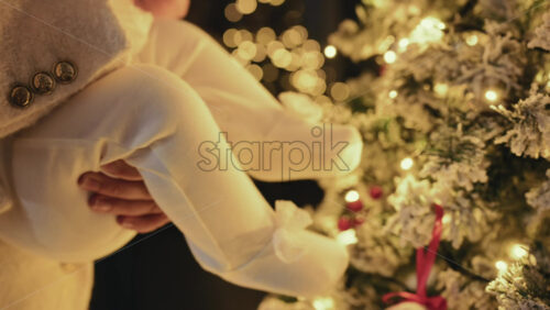 Video – A close, warmly lit shot of a baby dressed in a soft white outfit, being gently held by a mother’s hands - Starpik Stock