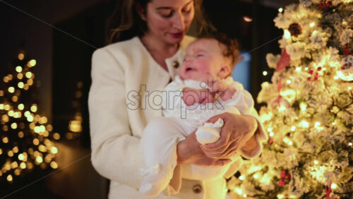 Video – A mother holding a baby, both dressed in white, while standing beside a decorated Christmas tree - Starpik Stock