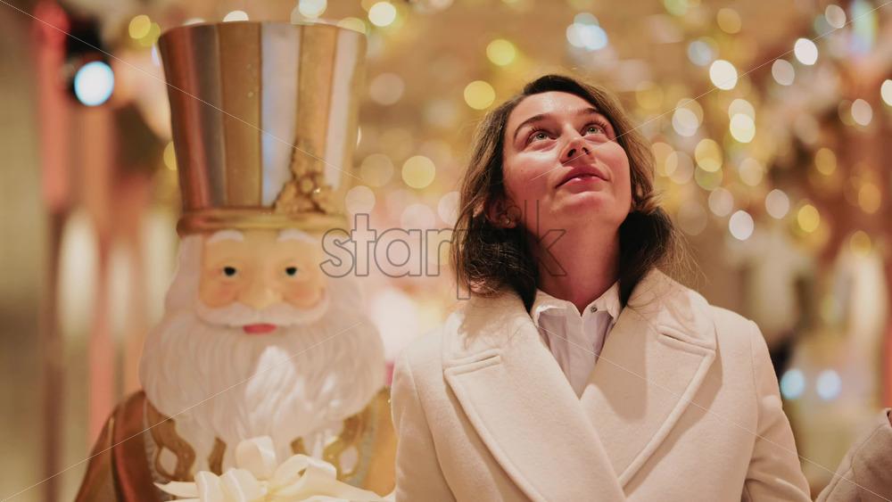 Video – A woman in a light colored coat stands beside a large nutcracker decoration, surrounded by warm festive lights - Starpik Stock