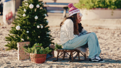 Video – Antibes, France – December 17, 2025: Young girls sitting on a small stool beside a decorated Christmas tree on a sandy beach taking pictures - Starpik Stock