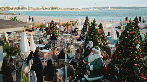 Video – Cannes, France – December 23, 2025: Wide view of a busy restaurant decorated for Christmas by the sea with decorated trees, holiday installations, and visitors - Starpik Stock