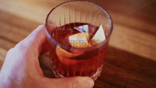 Video – Close up of a hand holding a glass filled with a red cocktail, ice cubes, and citrus peel - Starpik Stock