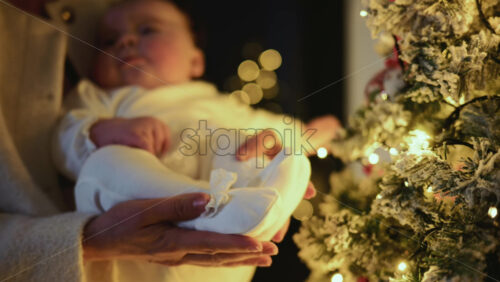 Video – Happy mother holding a baby indoors next to a decorated Christmas tree with warm lighting and soft background bokeh creating an intimate, festive atmosphere - Starpik Stock