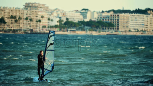 Video – Vallauris, France – December 27, 2025: Single windsurfer standing upright on a board while holding a sail in choppy water close to the coast - Starpik Stock