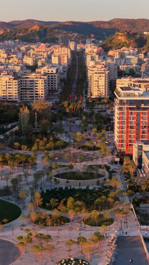 Video - Aerial drone view of the golden hour lighting highlighting Vlora's central urban park, featuring circular pathways, palm trees, playgrounds, and landscaped green spaces. Durres, Albania, Vertical
