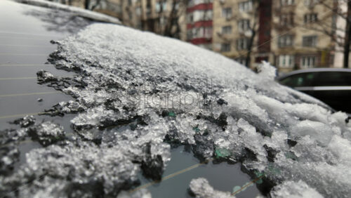 Close up of frozen ice crystals covering a car windshield, with urban buildings blurred in the background during cold winter conditions