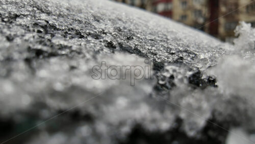 Close up of frozen ice crystals covering a car windshield, with urban buildings blurred in the background during cold winter conditions