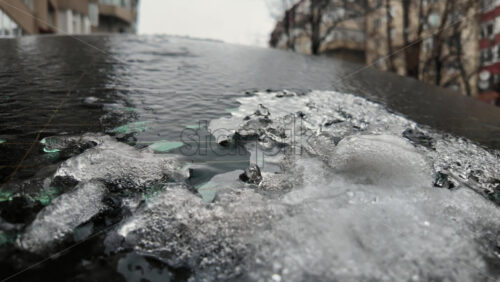 Close up of frozen ice crystals covering a car windshield, with urban buildings blurred in the background during cold winter conditions