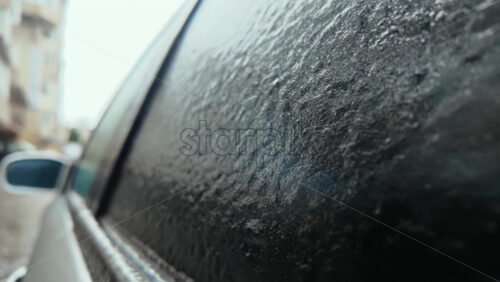 Side view of a car window covered in frost and ice, creating a textured frozen surface with urban background