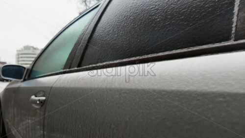 Side view of a car window covered in frost and ice, creating a textured frozen surface with urban background