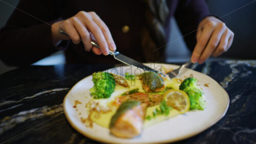 Close up of hands using knife and fork to cut a salmon fillet served with broccoli and sauce