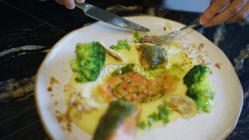 Close up of hands using knife and fork to cut a salmon fillet served with broccoli and sauce
