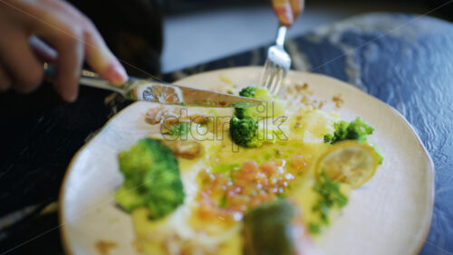 Close up of hands using knife and fork to cut a salmon fillet served with broccoli and sauce