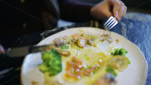 Close up of hands using knife and fork to cut a salmon fillet served with broccoli and sauce