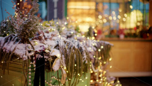 Close up of a decorative planter filled with winter greenery, covered in snow and wrapped with small glowing lights