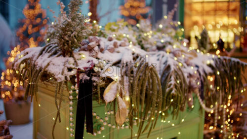 Close up of a decorative planter filled with winter greenery, covered in snow and wrapped with small glowing lights