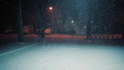 Snow falling steadily in an empty park at night. Streetlights glow softly in the background while snow covers the ground