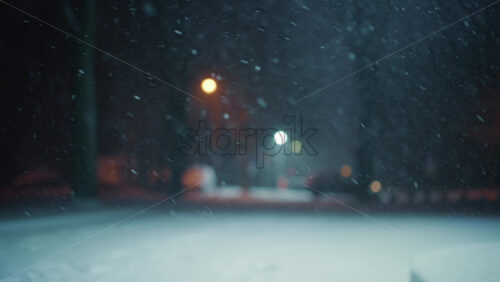 Snow falling steadily in an empty park at night. Streetlights glow softly in the background while snow covers the ground