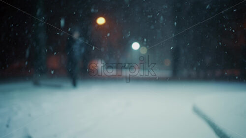 Snow falling steadily in an empty park at night. Streetlights glow softly in the background while snow covers the ground