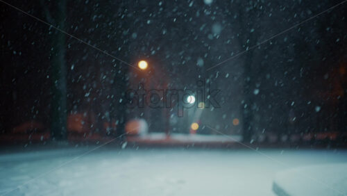 Snow falling steadily in an empty park at night. Streetlights glow softly in the background while snow covers the ground