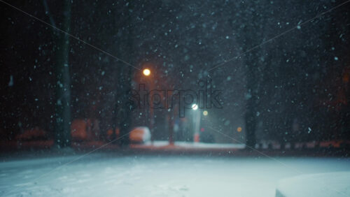 Snow falling steadily in an empty park at night. Streetlights glow softly in the background while snow covers the ground