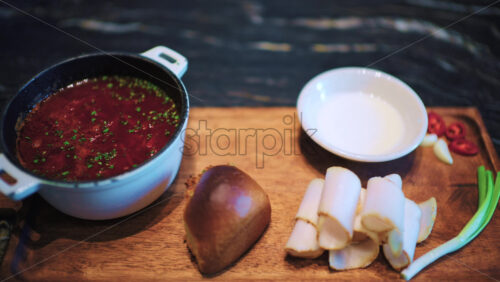 Close up of a bowl filled with borscht served on a wooden board alongside bread, sliced, cured pork fat, and spring onions