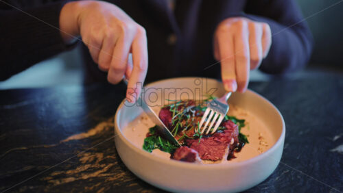 Close up of a woman's hands cutting and eating a plated steak with fork and knife