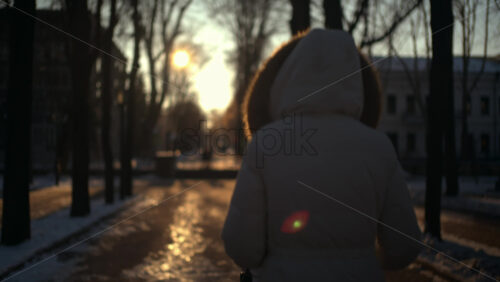 Rear view of a woman wearing a winter coat walking through a park at sunset, pushing a baby stroller