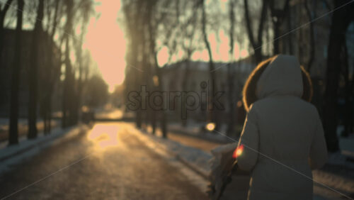 Rear view of a woman wearing a winter coat walking through a park at sunset, pushing a baby stroller