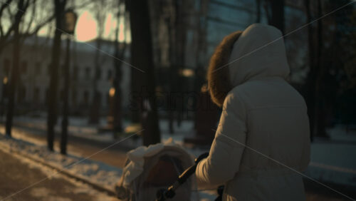 Rear view of a woman wearing a winter coat walking through a park at sunset, pushing a baby stroller