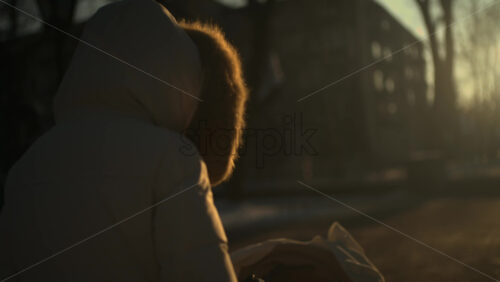 Rear view of a woman wearing a winter coat walking through a park at sunset, pushing a baby stroller