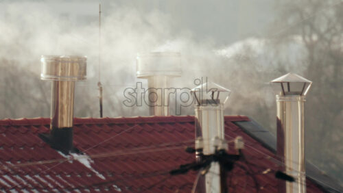 Close up of multiple metal chimneys releasing smoke above a red tiled residential roof on a cold day