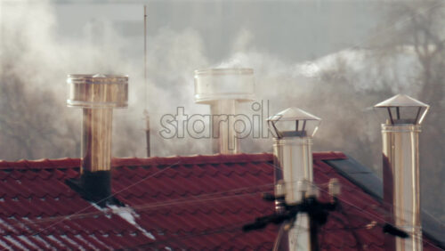 Close up of multiple metal chimneys releasing smoke above a red tiled residential roof on a cold day