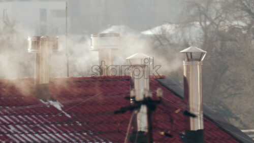 Close up of multiple metal chimneys releasing smoke above a red tiled residential roof on a cold day