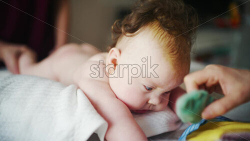 Infant lying on tummy on a soft pillow while reaching toward a small toy, supported by a caregiver doing mobility exercises