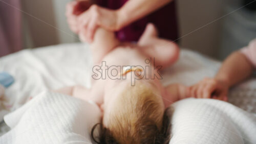 Infant lying relaxed on a bed, looking upward while a caregiver and the mother stay nearby doing mobility exercises