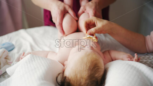 Infant lying relaxed on a bed, looking upward while a caregiver and the mother stay nearby doing mobility exercises