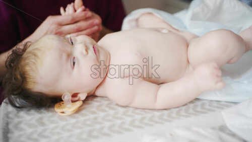 Infant lying relaxed on a bed, looking upward while a caregiver and the mother stay nearby doing mobility exercises