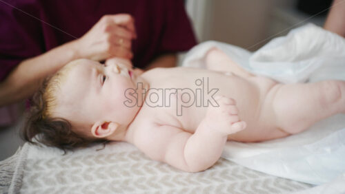 Infant lying relaxed on a bed, looking upward while a caregiver and the mother stay nearby doing mobility exercises