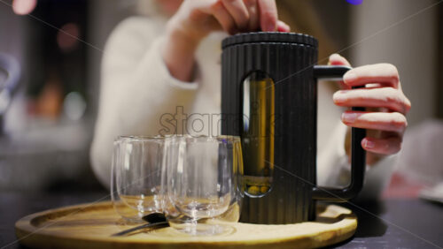 Hands preparing hot tea using a modern French press on a wooden tray with glass cups