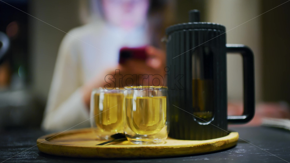 Two cups of freshly prepared tea on a tray while a woman uses her smartphone in the background