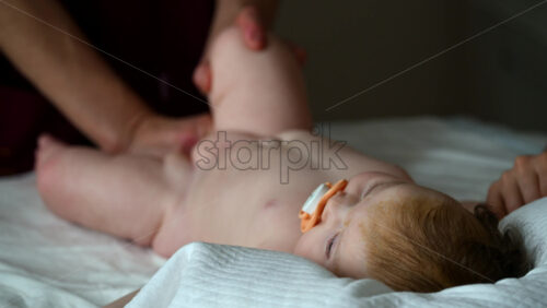 Infant lying relaxed on a bed, looking upward while a caregiver and the mother stay nearby doing mobility exercises