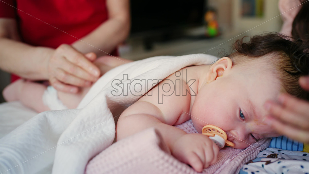 Infant resting on a soft blanket with a pacifier, gently supported by an adult's hand