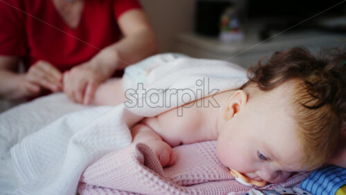 Close up of baby lying on tummy with blanket draped over the back, pacifier in mouth, and caregiver nearby